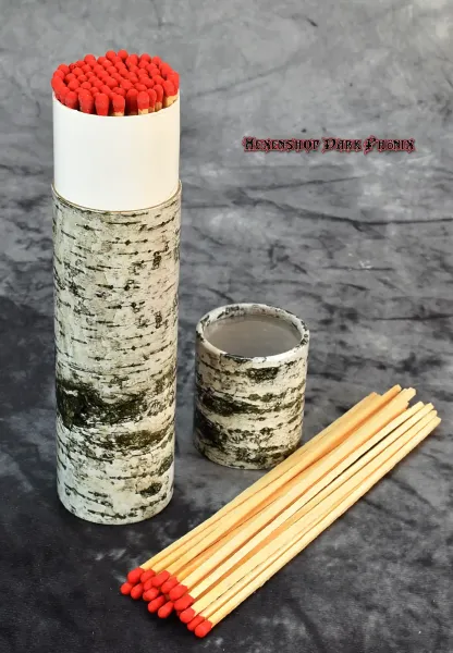Hexenshop Dark Phoenix Ein 20 cm langes Streichholz entzündet eine tiefe Ritualglaskerze, dekorative Baumrinden-Verpackung im Hintergrund.