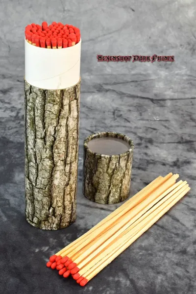 Hexenshop Dark Phoenix Ein 20 cm langes Streichholz entzündet eine tiefe Ritualglaskerze, dekorative Baumrinden-Verpackung im Hintergrund.